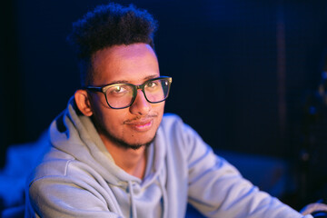 Obraz premium Dark skinned young man with a beaming smile, wears a gray parka, and glasses on dark background splashed by blue light