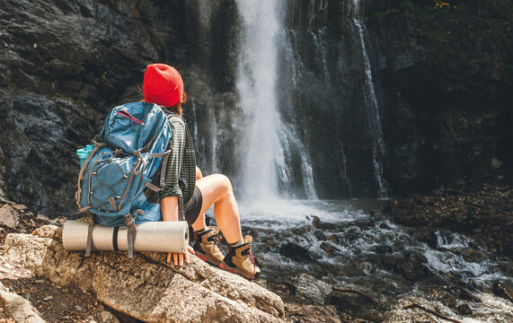 Woman with a backpack in a red hat dressed in active trekking clothes sitting near the mountain river waterfall and enjoying the splashing Nature power. Traveling, trekking, and a nature concept image