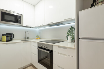 Fototapeta premium Kitchenette with plain white cabinets and chest of drawers, matching stone countertops and built-in appliances and white fridge