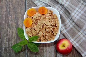 dry diet wheat muesli in a white plate, decorated with orange dried apricots and red apple