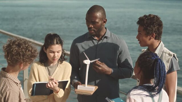 Medium slowmo of group of diverse elementary schoolchildren studying alternative energies outdoors listening to their modern African American male teacher holding windmill model