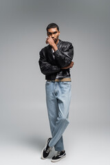 full length of stylish african american man in jeans and leather jacket posing on grey.