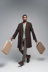 full length of stylish african american man in woolen coat and sneakers posing with shopping bags on grey.
