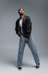 Fototapeta premium full length of stylish african american man in autumnal outfit posing with hand in pocket of jeans on grey.