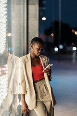 Portrait of a fashionable young woman with short hair and leaning her body against a shop window at night while using her phone