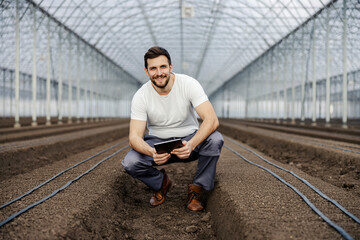 An agronomist assesses the quality of soil on the tablet while crouching at the greenhouse and smiling at the camera.