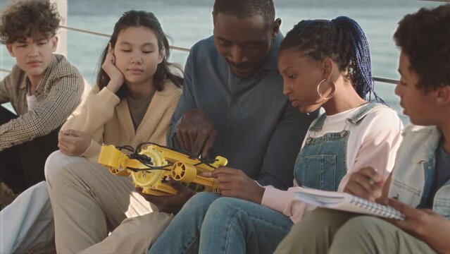 Medium slowmo of multiethnic school kids having outdoor engineering class with young male African American teacher, sitting at sunny quay and examining hydraulic centralizer