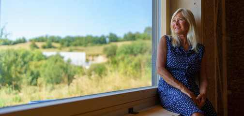Happy senior woman resting at window,enjoying view, inside of her new unfinished ecological wooden...
