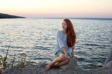 Fototapeta na wymiar A beautiful red-haired girl sits on a stone on the banks of a river, lake, sea. She is looking at ocean and thinking dreamily. Girl alone outside. Girl sitting on rocks. Lonely person.