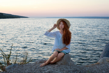 A beautiful red-haired girl in a hat sits on a stone on the banks of a river, lake, sea. The girl is alone on the street. The girl is sitting on the rocks. 