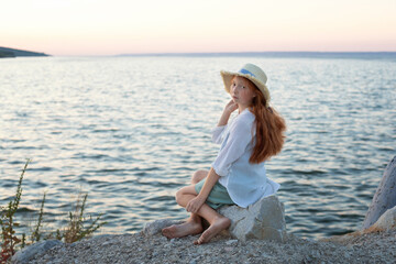 A beautiful red-haired girl in a hat sits on a stone on the banks of a river, lake, sea.