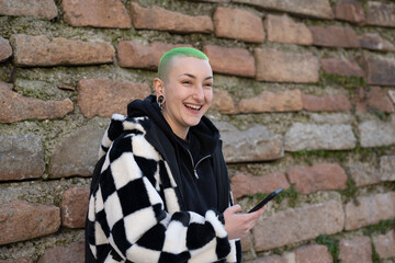 Fototapeta na wymiar portrait of young authentic androgynous woman with short haircut and colored green, genuine smiling lesbian woman using smartphone, happy girl with piercing and street fashion dress code