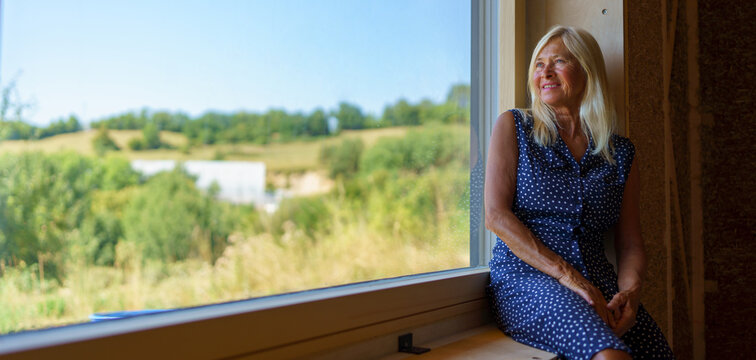 Happy senior woman resting at window,enjoying view, inside of her new unfinished ecological wooden house.