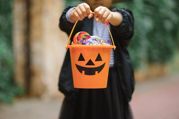 Little cute girl in witch costume holding jack-o-lantern pumpkin bucket with candies and sweets. Kid trick or treating in Halloween holiday. - obrazy, fototapety, plakaty