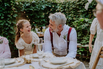 Happy groom talking and smiling with his little daughter during outdoor wedding party.