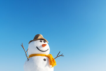 Funny snowman in stylish brown hat and yellow scalf on snowy field. Blue sky on background
