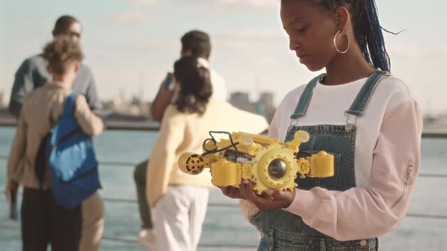 Medium slowmo portrait of African American schoolgirl smiling at camera while holding hydraulic centralizer, doing engineering school project with teacher and classmates outdoors at sunny quay