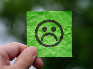 A man holding a green note paper with a sad face on it in his hand against a background of foliage