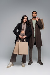 full length of fashionable interracial couple in stylish outfits holding shopping bags on grey.