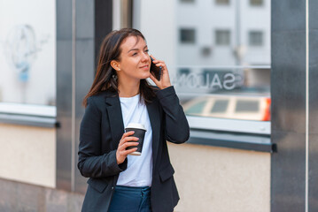 Business woman with coffe paper cup talking on the phone near office.Businesswoman wears a black suit and a white T-shirt.Office building blurred background.Coffe time concept.Summer day.