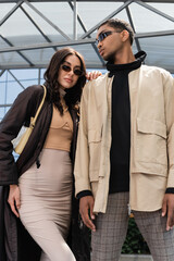 low angle view of stylish interracial couple in sunglasses and stylish outfits looking at camera.