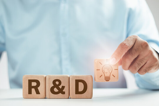 A man's hand holds a wooden block with a lightbulb icon and a wooden block with the word RD, signifying research and development, whether in education or business, to progress further. Brain icon.