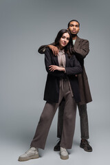 full length of african american man hugging brunette woman in blazer isolated on grey.
