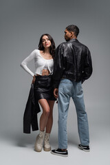 full length of woman in leather skirt holding black jacket near stylish african american man on grey.