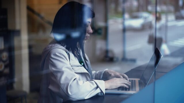 Young woman working remotely in front of laptop at  business office by window overlooking urban street sidewalk reflection
