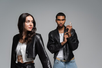 young and stylish multiethnic couple in black leather jackets posing isolated on grey.