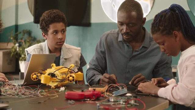 Waist up slowmo of African American school kids having electronics and robotics class at school with young Black man teaching and helping them