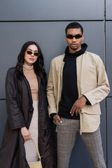 young multiethnic couple in trendy autumnal outfits and sunglasses posing near building.