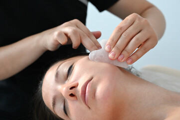 Fototapeta na wymiar Young woman receiving facial massage with gua sha tool in beauty salon. Close up. Beauty salon