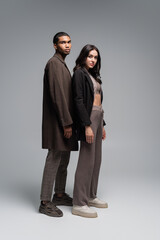 full length of stylish interracial couple in autumnal outfits standing together on grey.