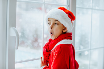 Boy in red Santa Claus suit posing near big window with snowing outside.