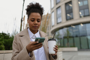 african american young female student looking at a mobile phone with a cup of coffee on the background of the city