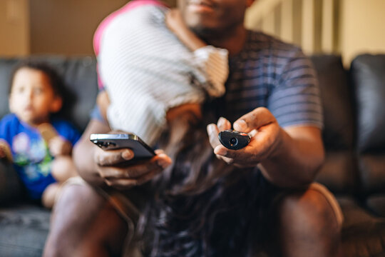 Father At Home Sitting On Couch With Kids In The Background While Holding Tv Remote And Cellphone