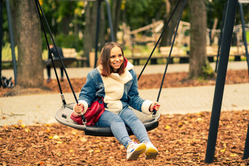 A beautiful and cheerful woman in warm clothes swings on a swing in the park. Girl swinging in the autumn city park