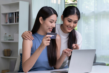 Young Asian lesbian couple holding credit card and using laptop computer. LGBT lesbian businesswoman working at home. Online shopping, e-commerce, internet banking, spending money, working from home 