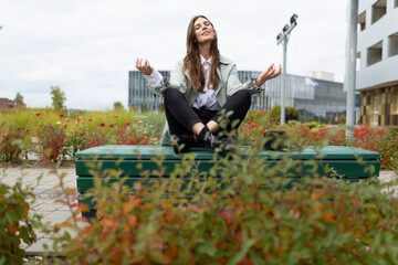 a young woman sits on a bench in the park in a yoga pose with her arms apart with her eyes closed