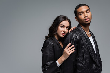 young woman in leather jacket hugging stylish african american man isolated on grey.