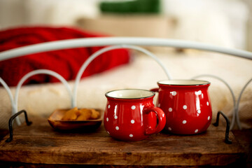 Two red mugs with white dots on a tray with a plate with cookies in front of a bed