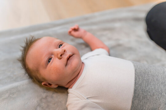 White blond infant boy in white t-shirt lying on his back on blanket on the floor looking at his mother out of shot with curious expression. Bonding time. Horizontal indoor shot. High quality photo