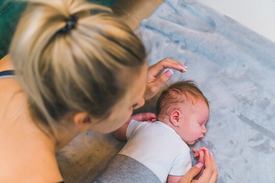 Caucasian blond woman leaning over her newborn baby boy lying on blanket on the floor next to couch. Caring mother. Responsibility. Horizontal indoor shot. High quality photo