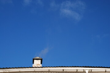 Chimney against the blue sky. The white brick chimney of the spherhu stove is covered with an iron umbrella deflector to protect it from moisture. Barely noticeable gray smoke comes out of the chimney