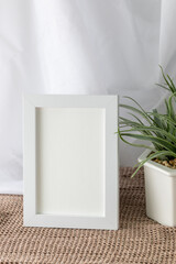 Blank vertical white frame beside a houseplant, painting or artwork display on brown knitted cloth