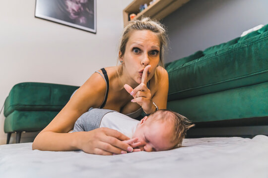 Closeup indoor shot of a caucasian middle-aged mother shushing the camera by covering her lips with index finger in order to remind everyone that her adorable infant baby boy is sleeping deeply on