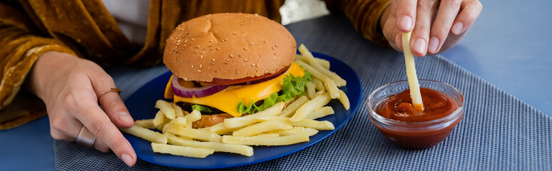 partial view of woman dipping french fries into tomato sauce near delicious burger on blue plate, banner.