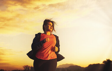 A beautiful curvy young adult woman jogger runs in a nature at sunset - determination and weight...
