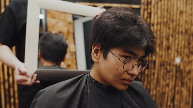 Oriental young man with glasses, looking at his hairstyle in mirror, sitting in a barbershop chair assessing hairstyle. Barber holds a mirror, showing client the waves from the back